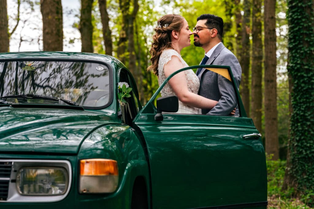 Mooi bruidspaar dat een romantisch moment deelt in een weelderige bosomgeving met een oldtimer groene wagen